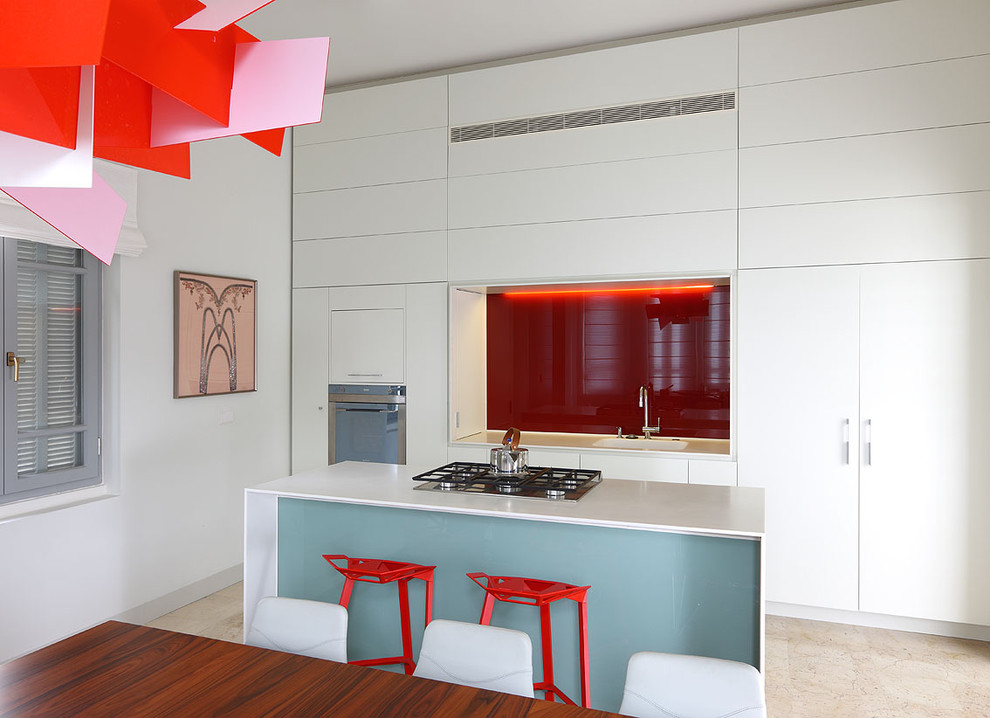 Diseño de cocina comedor moderna pequeña con armarios con paneles lisos, puertas de armario blancas, encimera de acrílico, salpicadero rojo, salpicadero de vidrio templado, electrodomésticos de acero inoxidable, fregadero encastrado, suelo de piedra caliza y una isla