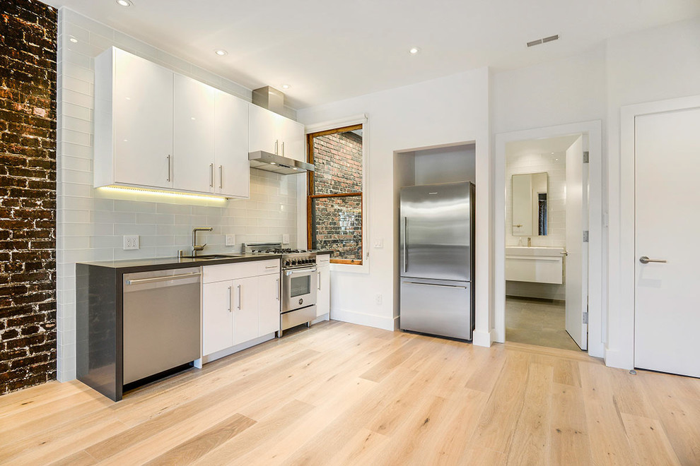 Modern inredning av ett litet linjärt kök med öppen planlösning, med en undermonterad diskho, vita skåp, bänkskiva i betong, vitt stänkskydd, stänkskydd i glaskakel, rostfria vitvaror, ljust trägolv och luckor med upphöjd panel