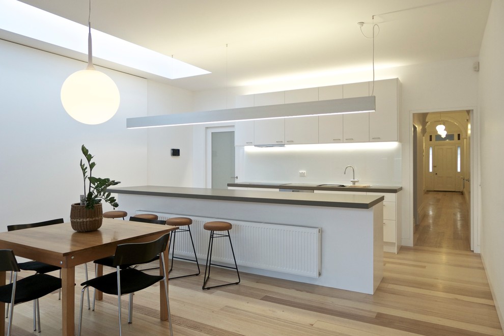 Offene, Zweizeilige Moderne Küche mit flächenbündigen Schrankfronten, weißen Schränken, Kücheninsel, Quarzwerkstein-Arbeitsplatte, Küchenrückwand in Weiß, Glasrückwand und braunem Holzboden in Melbourne