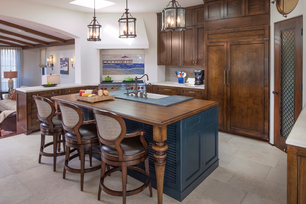 Enclosed kitchen - mediterranean galley travertine floor and beige floor enclosed kitchen idea in Los Angeles with a farmhouse sink, recessed-panel cabinets, dark wood cabinets, wood countertops, multicolored backsplash, mosaic tile backsplash, paneled appliances and an island