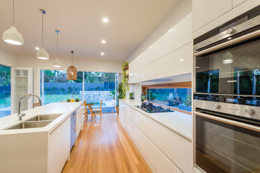 Moderne Wohnküche mit Doppelwaschbecken, flächenbündigen Schrankfronten, weißen Schränken, Küchengeräten aus Edelstahl, hellem Holzboden und Kücheninsel in Brisbane