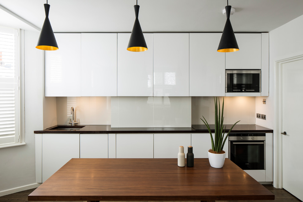 Exempel på ett litet modernt linjärt kök och matrum, med en undermonterad diskho, släta luckor, vita skåp, svarta vitvaror, mörkt trägolv, grått stänkskydd och glaspanel som stänkskydd