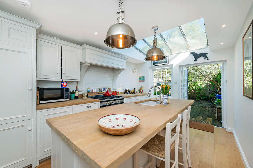 Photo of a medium sized farmhouse single-wall enclosed kitchen in London with a double-bowl sink, shaker cabinets, light wood cabinets, wood worktops, white splashback, metro tiled splashback, stainless steel appliances, light hardwood flooring, an island, beige floors and beige worktops.