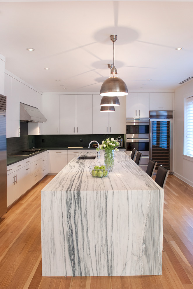 Inredning av ett modernt kök och matrum, med vita skåp, marmorbänkskiva, grönt stänkskydd, stänkskydd i sten och rostfria vitvaror