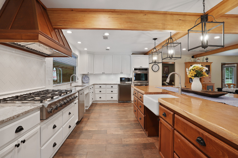 Inspiration for a large rustic u-shaped open plan kitchen in New Orleans with a belfast sink, white cabinets, wood worktops, white splashback, metro tiled splashback, stainless steel appliances, porcelain flooring, an island, brown floors, multicoloured worktops and raised-panel cabinets.