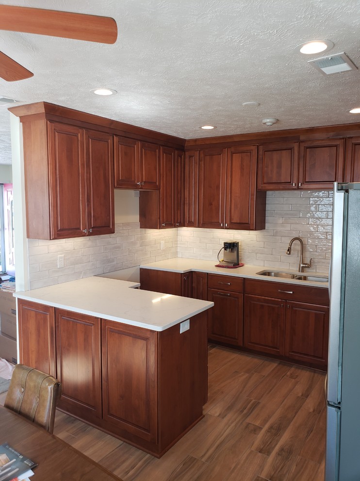 Klassisk inredning av ett mellanstort vit vitt kök, med en dubbel diskho, luckor med upphöjd panel, skåp i mellenmörkt trä, bänkskiva i kvarts, vitt stänkskydd, stänkskydd i keramik, rostfria vitvaror, klinkergolv i keramik, en halv köksö och brunt golv