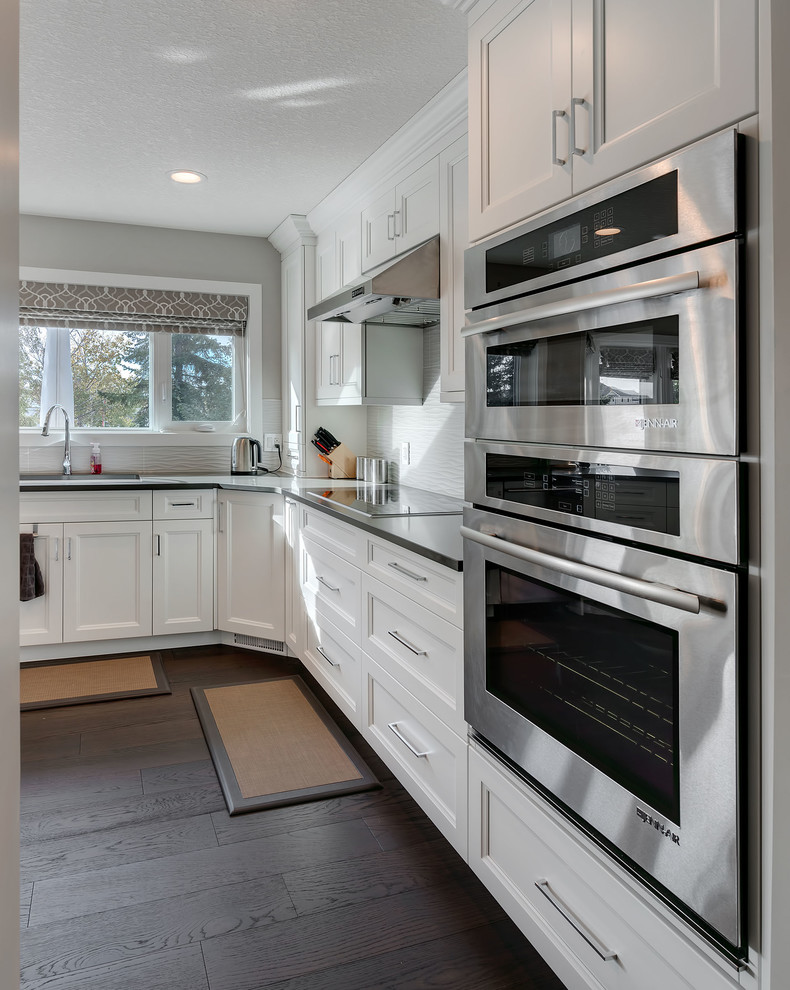 Medium sized retro l-shaped kitchen/diner in Vancouver with a submerged sink, shaker cabinets, white cabinets, grey splashback, black appliances, dark hardwood flooring and a breakfast bar.
