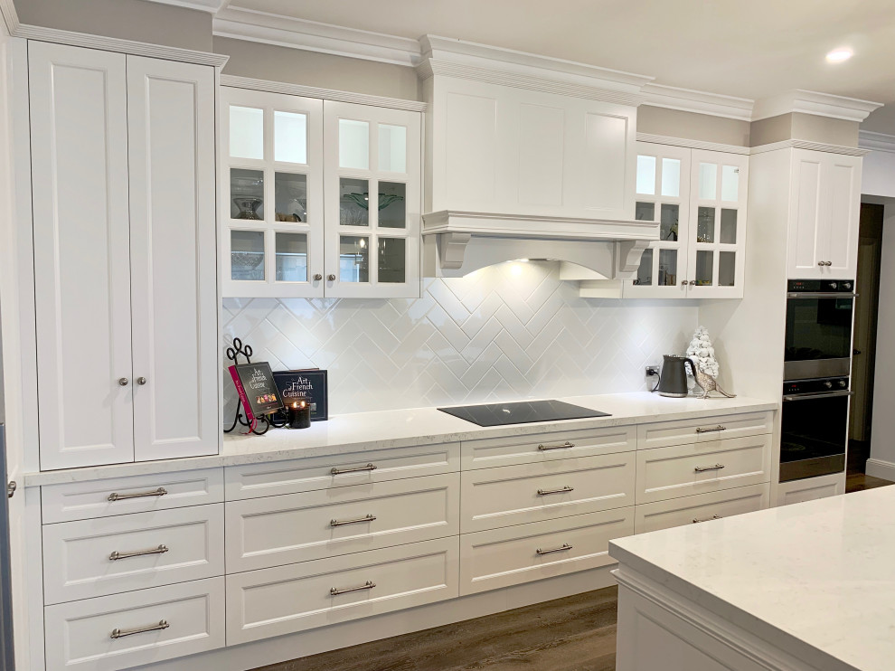 Foto på ett mycket stort vintage vit kök, med en undermonterad diskho, luckor med infälld panel, grå skåp, bänkskiva i kvarts, vitt stänkskydd, stänkskydd i sten, svarta vitvaror, mellanmörkt trägolv, en köksö och brunt golv