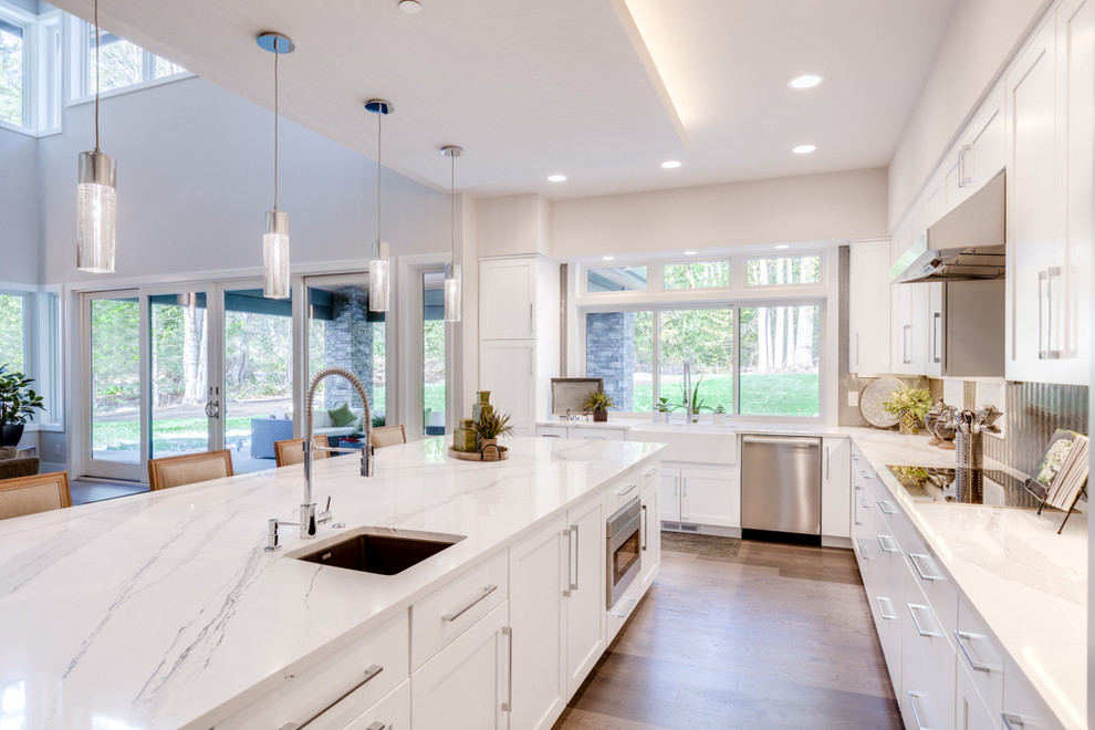 Medium sized contemporary l-shaped open plan kitchen in Seattle with a belfast sink, shaker cabinets, white cabinets, engineered stone countertops, metallic splashback, integrated appliances, light hardwood flooring, an island, beige floors and white worktops.