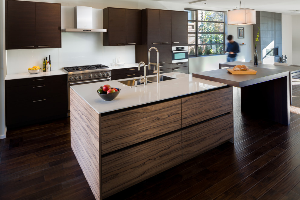 Inspiration for a large modern single-wall kitchen/diner in Seattle with engineered stone countertops, dark hardwood flooring, brown floors, white worktops, a submerged sink, flat-panel cabinets, dark wood cabinets, stainless steel appliances, an island, white splashback and glass tiled splashback.