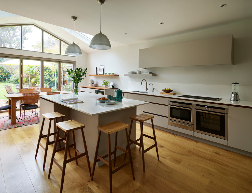 Large contemporary l-shaped kitchen/diner in London with flat-panel cabinets, beige cabinets, composite countertops, medium hardwood flooring, an island, beige floors and white worktops.