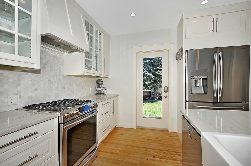 Example of a mid-sized transitional u-shaped light wood floor open concept kitchen design in Calgary with a farmhouse sink, raised-panel cabinets, white cabinets, marble countertops, stainless steel appliances and an island