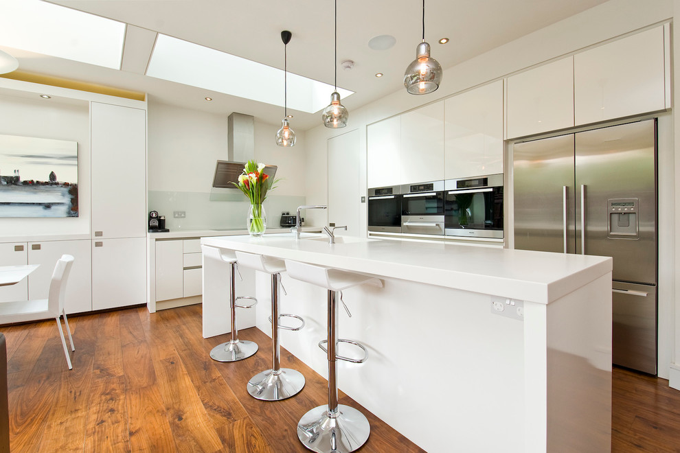 Moderne Wohnküche in L-Form mit flächenbündigen Schrankfronten, weißen Schränken, Küchengeräten aus Edelstahl, braunem Holzboden und Kücheninsel in London