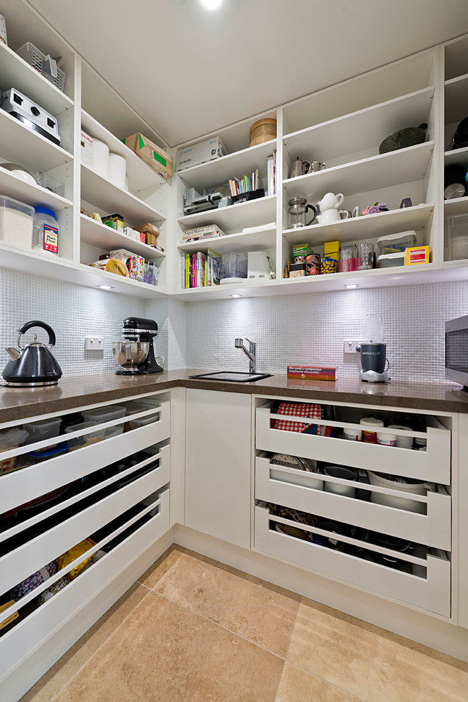 Modern inredning av ett kök, med en undermonterad diskho, släta luckor, vita skåp, bänkskiva i kvarts, rostfria vitvaror, en köksö, vitt stänkskydd, stänkskydd i mosaik och travertin golv