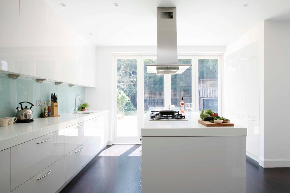 Large modern galley kitchen/diner in London with a submerged sink, flat-panel cabinets, white cabinets, composite countertops, white splashback, glass sheet splashback, stainless steel appliances, dark hardwood flooring and an island.