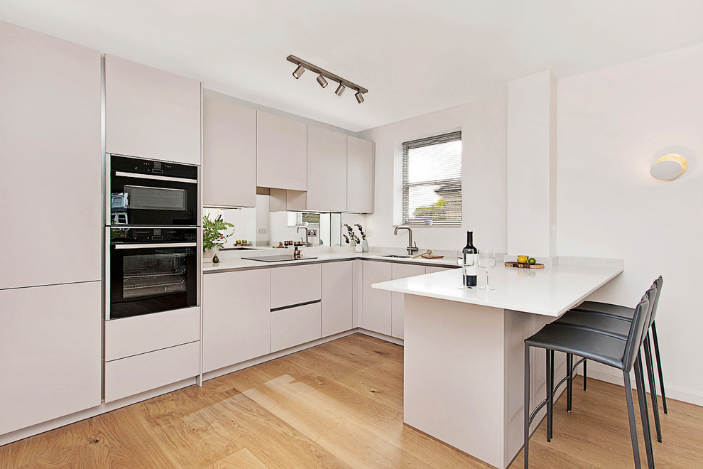 ロンドンにある高級な小さなモダンスタイルのおしゃれなキッチン (一体型シンク、フラットパネル扉のキャビネット、ベージュのキャビネット、クオーツストーンカウンター、白いキッチンパネル、ミラータイルのキッチンパネル、黒い調理設備、無垢フローリング、白いキッチンカウンター) の写真