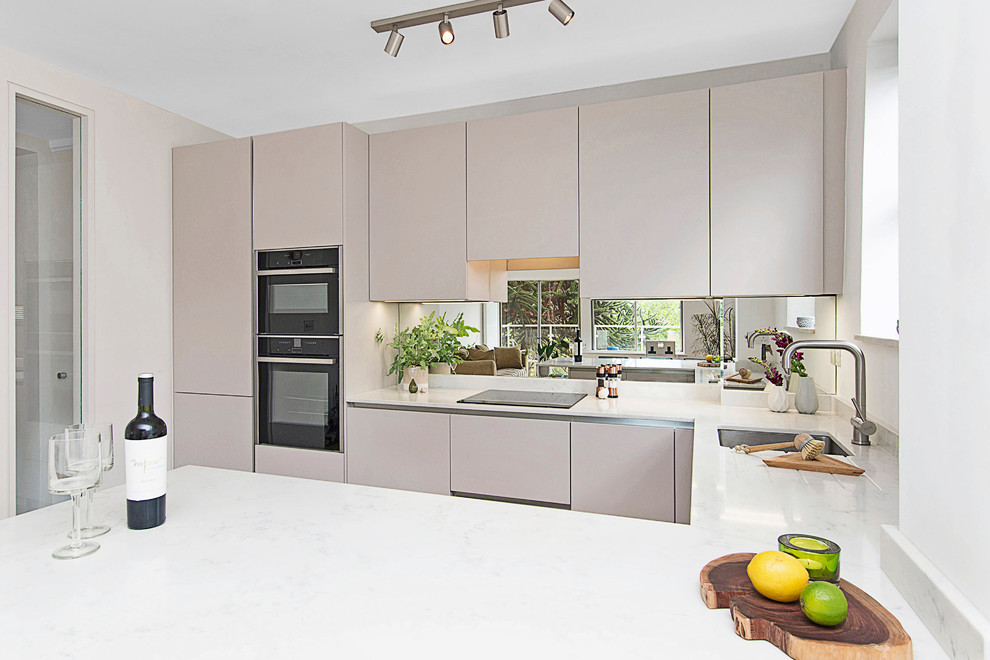 Photo of a small modern u-shaped kitchen/diner in London with an integrated sink, flat-panel cabinets, beige cabinets, engineered stone countertops, white splashback, mirror splashback, black appliances, medium hardwood flooring, a breakfast bar and white worktops.