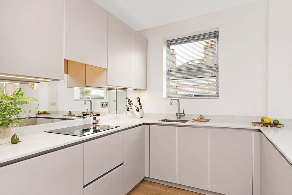 This is an example of a medium sized contemporary u-shaped kitchen in London with an integrated sink, flat-panel cabinets, beige cabinets, engineered stone countertops, mirror splashback, black appliances, medium hardwood flooring, a breakfast bar, beige floors and white worktops.
