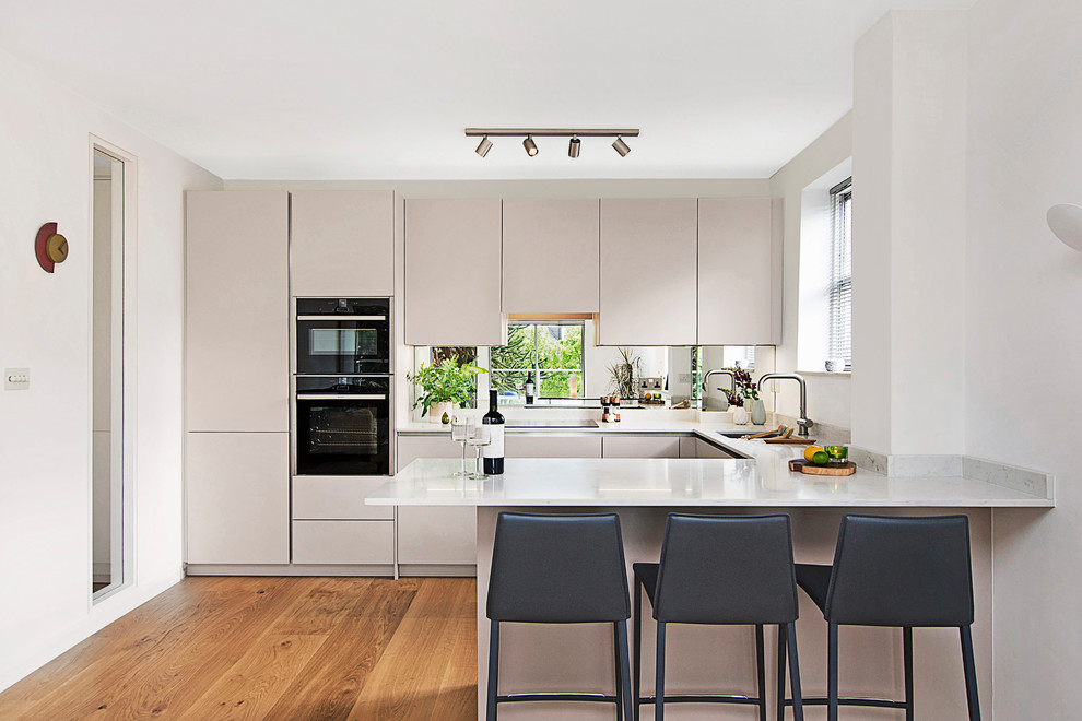 Inspiration for a medium sized contemporary u-shaped kitchen in London with flat-panel cabinets, beige cabinets, engineered stone countertops, mirror splashback, black appliances, medium hardwood flooring, a breakfast bar, white worktops and a submerged sink.