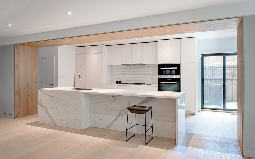 Design ideas for a large modern kitchen in Toronto with a submerged sink, flat-panel cabinets, white cabinets, white splashback, metro tiled splashback, integrated appliances, light hardwood flooring, an island and brown floors.