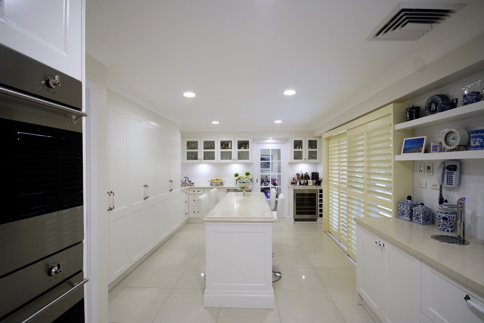 Design ideas for a large classic u-shaped open plan kitchen in Sydney with a belfast sink, recessed-panel cabinets, white cabinets, engineered stone countertops, white splashback, metro tiled splashback, stainless steel appliances, ceramic flooring, an island, beige floors and beige worktops.