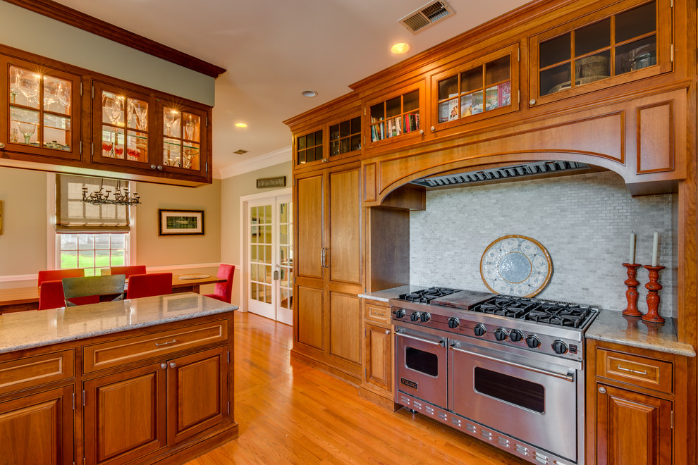 Large elegant u-shaped light wood floor eat-in kitchen photo in New York with a farmhouse sink, glass-front cabinets, medium tone wood cabinets, granite countertops, gray backsplash, stone tile backsplash and paneled appliances