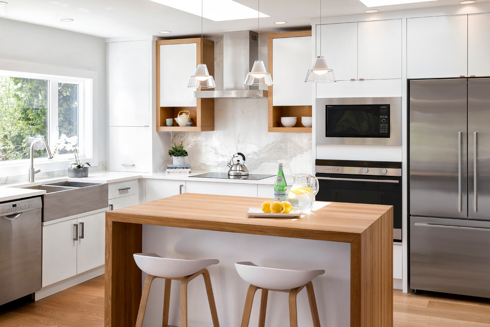 Scandi l-shaped kitchen in Vancouver with a belfast sink, flat-panel cabinets, white cabinets, white splashback, stainless steel appliances, light hardwood flooring, an island and beige floors.