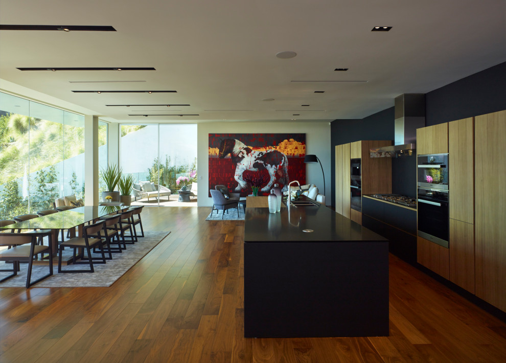 This is an example of a large modern galley kitchen/diner in Los Angeles with a double-bowl sink, flat-panel cabinets, black cabinets, glass worktops, black splashback, metal splashback, black appliances, dark hardwood flooring, an island and black worktops.
