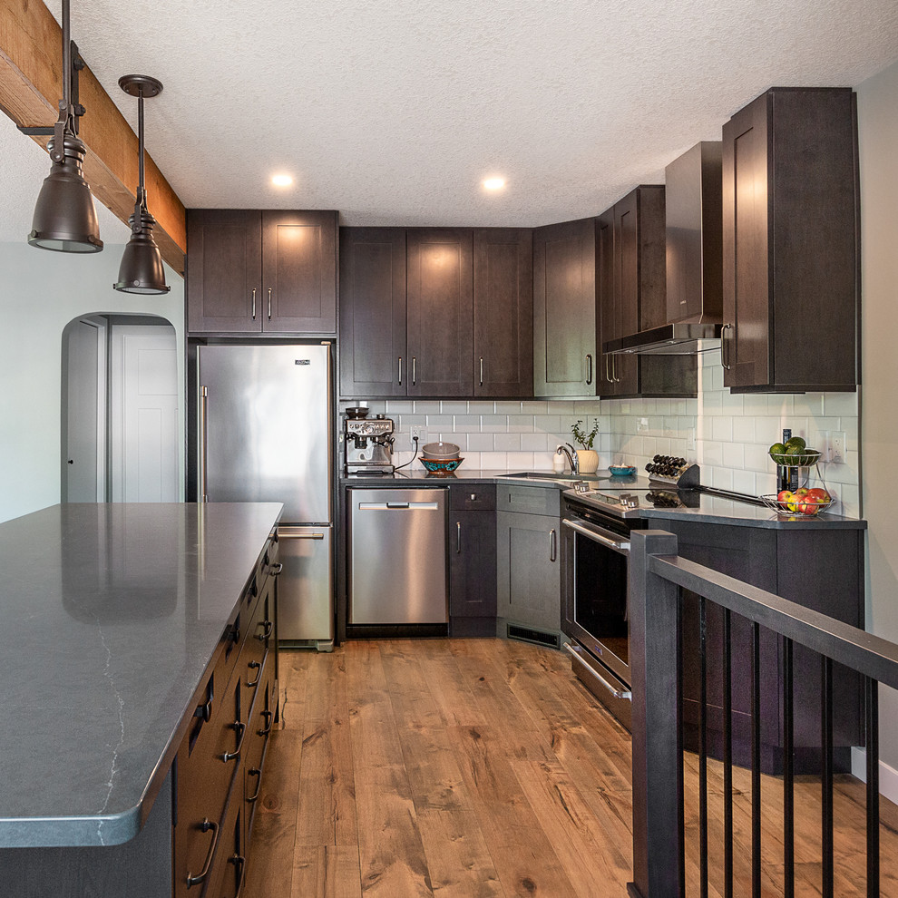 Bild på ett litet industriellt grå grått kök, med en undermonterad diskho, luckor med infälld panel, bruna skåp, bänkskiva i kvartsit, vitt stänkskydd, stänkskydd i keramik, rostfria vitvaror, mellanmörkt trägolv, en köksö och brunt golv