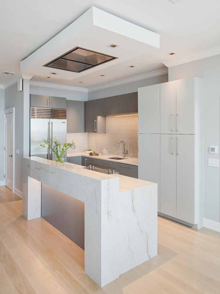 Design ideas for a contemporary u-shaped kitchen in Philadelphia with a submerged sink, flat-panel cabinets, grey cabinets, grey splashback, glass tiled splashback, stainless steel appliances, light hardwood flooring, a breakfast bar, beige floors and white worktops.