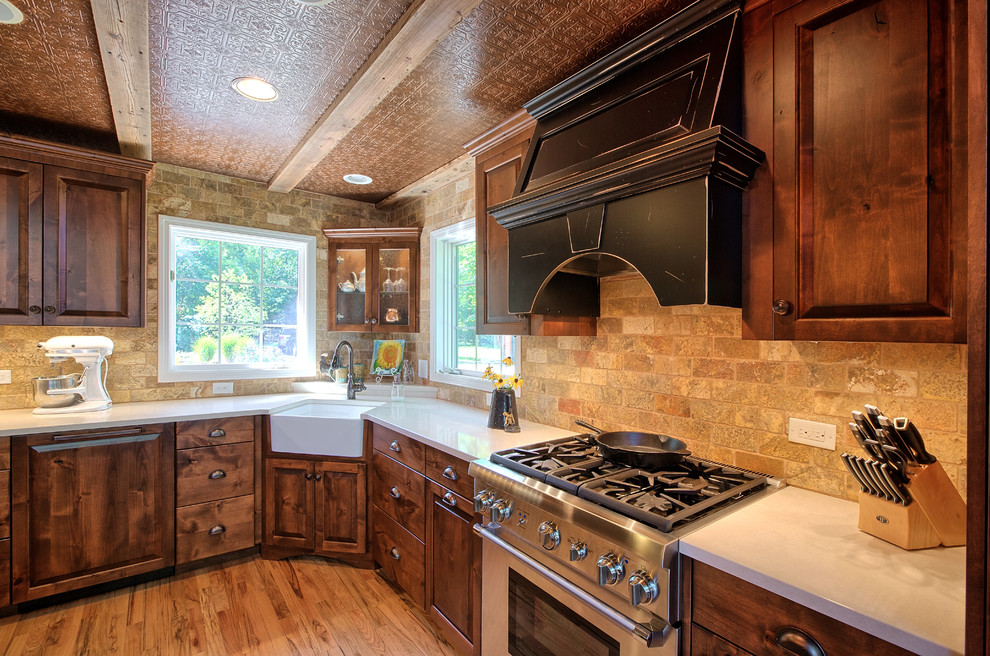 Inspiration for a medium sized rustic l-shaped kitchen/diner in Minneapolis with a belfast sink, raised-panel cabinets, dark wood cabinets, engineered stone countertops, multi-coloured splashback, travertine splashback, stainless steel appliances, light hardwood flooring, an island and multi-coloured floors.