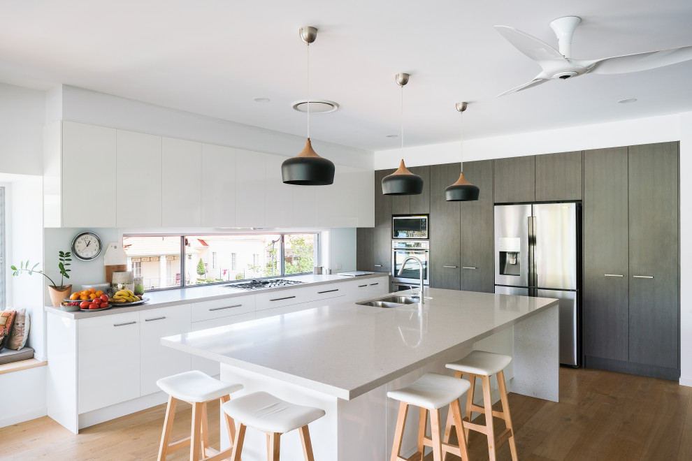Design ideas for a medium sized contemporary l-shaped open plan kitchen in Brisbane with a submerged sink, flat-panel cabinets, white cabinets, engineered stone countertops, stainless steel appliances, an island, white worktops, window splashback, medium hardwood flooring and brown floors.