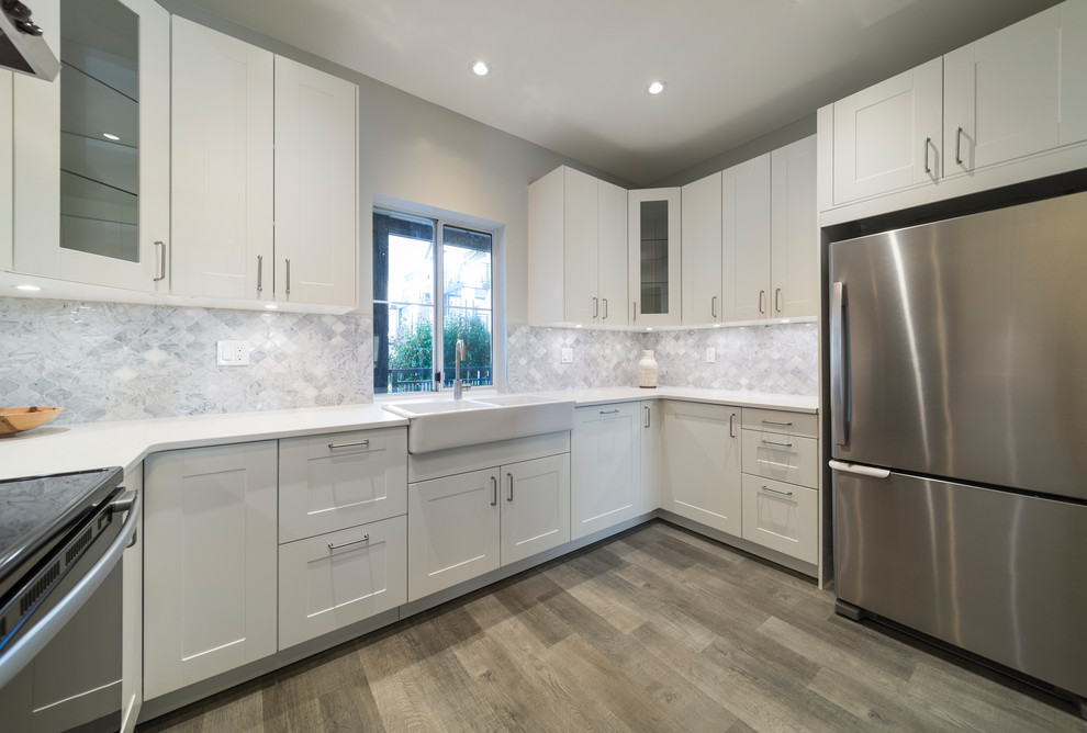 This is an example of a medium sized contemporary u-shaped enclosed kitchen in Vancouver with a belfast sink, shaker cabinets, white cabinets, engineered stone countertops, grey splashback, marble splashback, stainless steel appliances, laminate floors, no island, brown floors and white worktops.