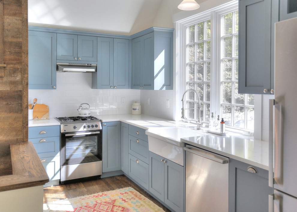 Inredning av ett lantligt mellanstort vit vitt kök, med en rustik diskho, skåp i shakerstil, blå skåp, bänkskiva i kvarts, vitt stänkskydd, stänkskydd i porslinskakel, rostfria vitvaror, mellanmörkt trägolv och brunt golv