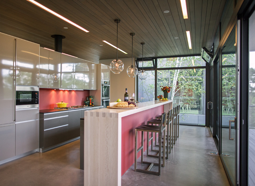 Trendy galley concrete floor eat-in kitchen photo in New York with a drop-in sink, glass-front cabinets, beige cabinets, granite countertops, red backsplash, glass sheet backsplash, stainless steel appliances and an island