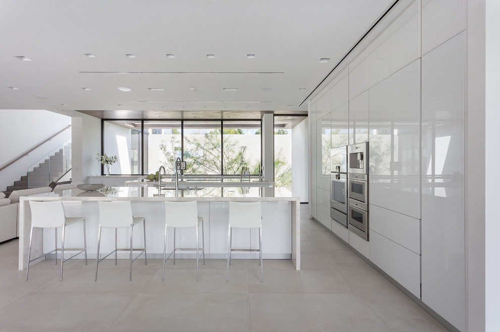 Свежая идея для дизайна: кухня-гостиная в стиле модернизм с врезной мойкой, плоскими фасадами, белыми фасадами, техникой из нержавеющей стали, бежевым полом и двумя и более островами - отличное фото интерьера