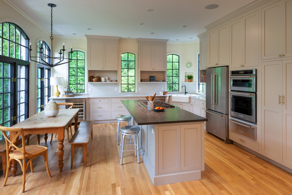 Example of a cottage l-shaped medium tone wood floor and brown floor eat-in kitchen design in Richmond with a farmhouse sink, shaker cabinets, beige cabinets, white backsplash, stainless steel appliances, an island and white countertops