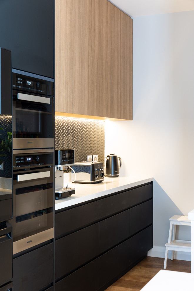 Photo of a large contemporary kitchen pantry in Sydney with a built-in sink, flat-panel cabinets, engineered stone countertops, black splashback, mosaic tiled splashback, black appliances, light hardwood flooring, an island and white worktops.