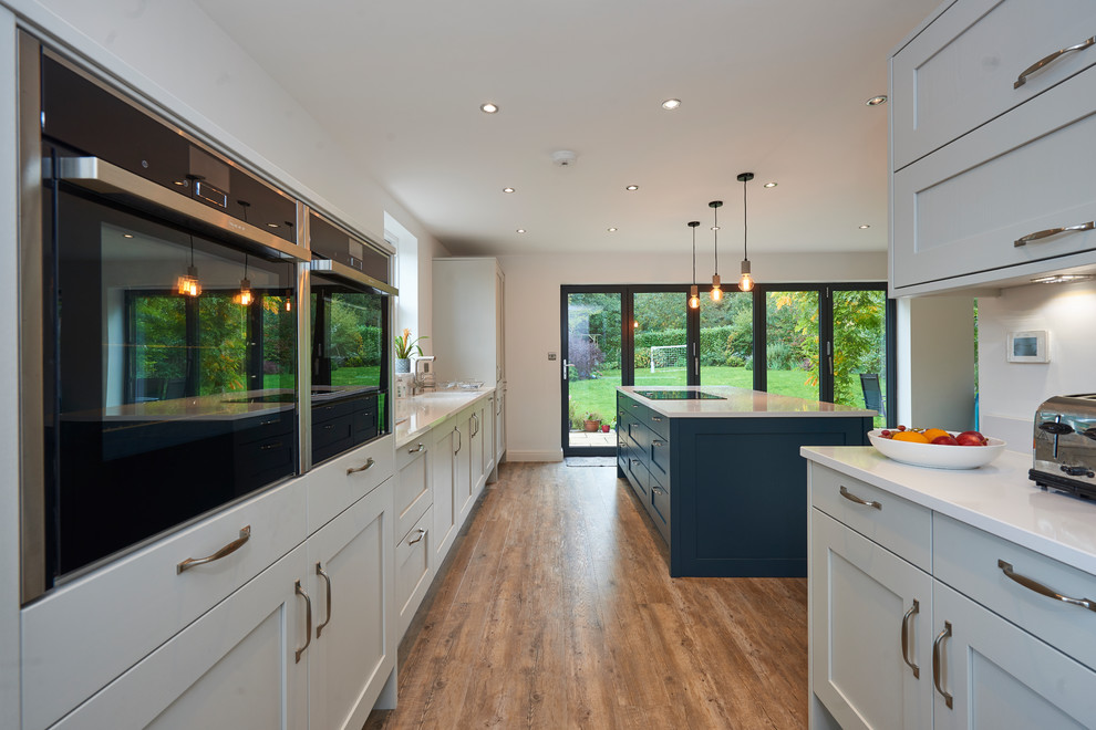 Example of a transitional galley medium tone wood floor and brown floor eat-in kitchen design in Other with shaker cabinets, gray cabinets, black appliances, an island and white countertops