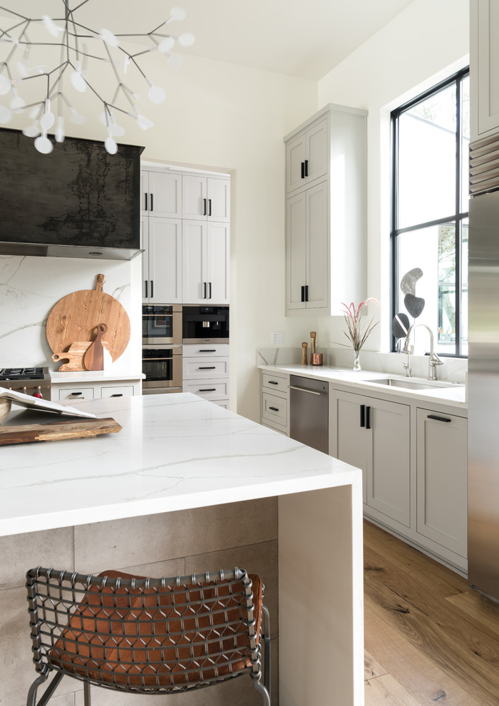 Exemple d'une cuisine chic en L avec un évier encastré, un placard à porte shaker, des portes de placard grises, un électroménager en acier inoxydable, parquet clair, îlot, un sol beige et un plan de travail blanc.