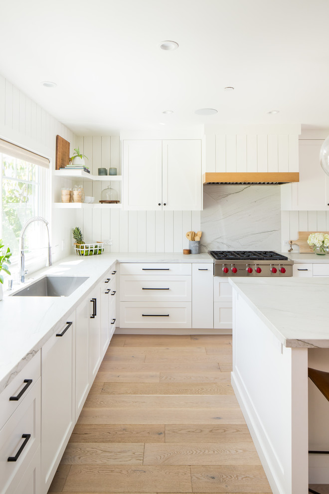 This is an example of a traditional l-shaped kitchen/diner in Orange County with a submerged sink, shaker cabinets, white cabinets, quartz worktops, white splashback, wood splashback, integrated appliances, light hardwood flooring, an island and white worktops.
