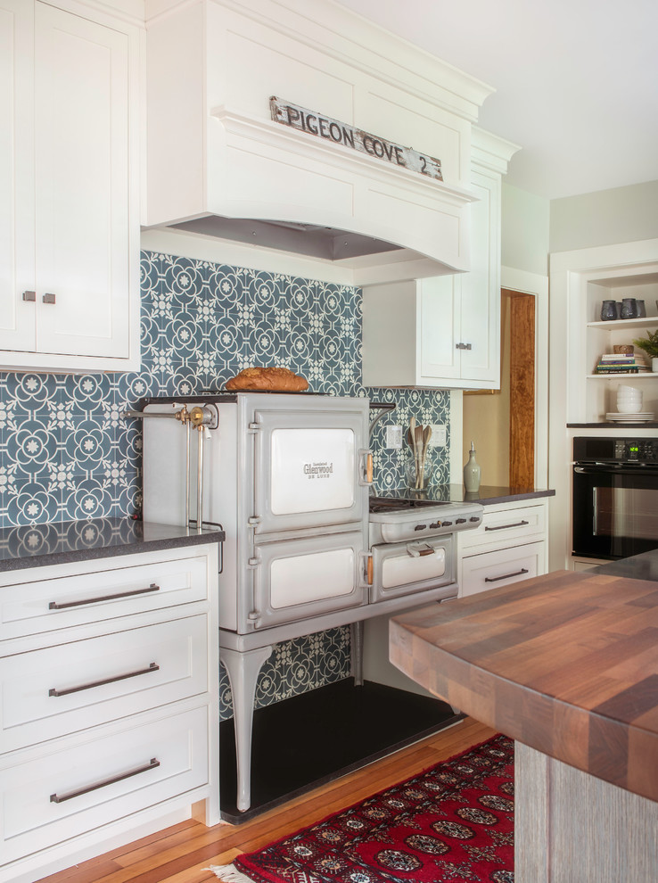 Example of a mid-sized mountain style galley medium tone wood floor and brown floor open concept kitchen design in Boston with a farmhouse sink, beaded inset cabinets, white cabinets, wood countertops, blue backsplash, cement tile backsplash, paneled appliances, an island and black countertops