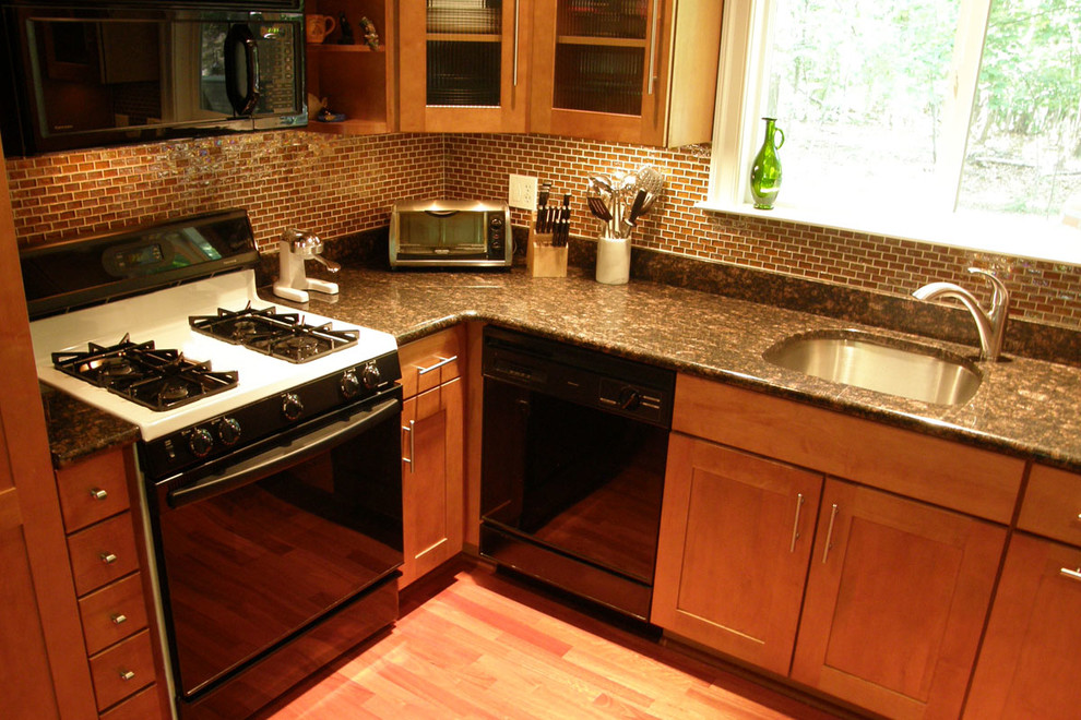 Foto på ett avskilt, mellanstort funkis l-kök, med en undermonterad diskho, luckor med infälld panel, skåp i mellenmörkt trä, granitbänkskiva, orange stänkskydd, stänkskydd i glaskakel, svarta vitvaror och mellanmörkt trägolv
