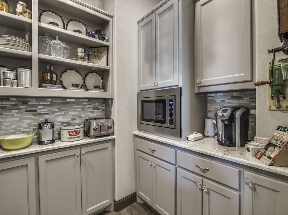 This is an example of a large traditional kitchen pantry in Dallas with a belfast sink, recessed-panel cabinets, grey cabinets, granite worktops, grey splashback, glass tiled splashback, stainless steel appliances, dark hardwood flooring, multiple islands and brown floors.