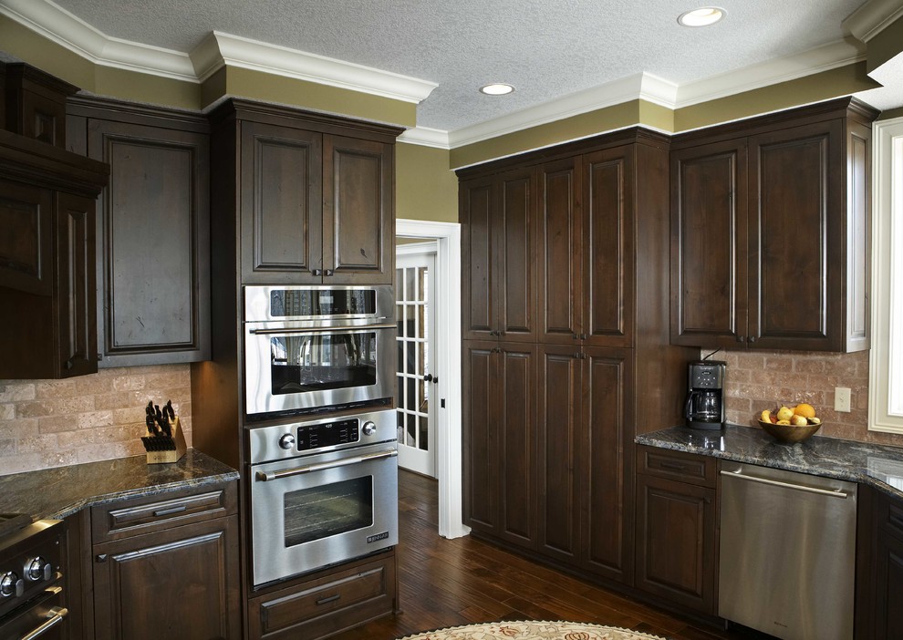 Example of a classic eat-in kitchen design in Minneapolis with an undermount sink, raised-panel cabinets, dark wood cabinets, granite countertops, beige backsplash, stone tile backsplash and stainless steel appliances