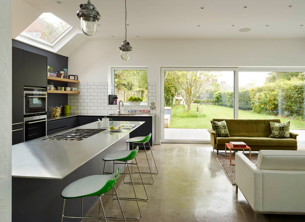 Large contemporary l-shaped open plan kitchen in London with an integrated sink, flat-panel cabinets, black cabinets, stainless steel worktops, white splashback, ceramic splashback, concrete flooring, no island and beige floors.