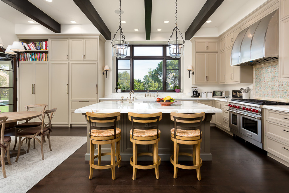 Photo of a mediterranean l-shaped kitchen in Los Angeles with a belfast sink, recessed-panel cabinets, beige cabinets, multi-coloured splashback, stainless steel appliances, dark hardwood flooring and an island.