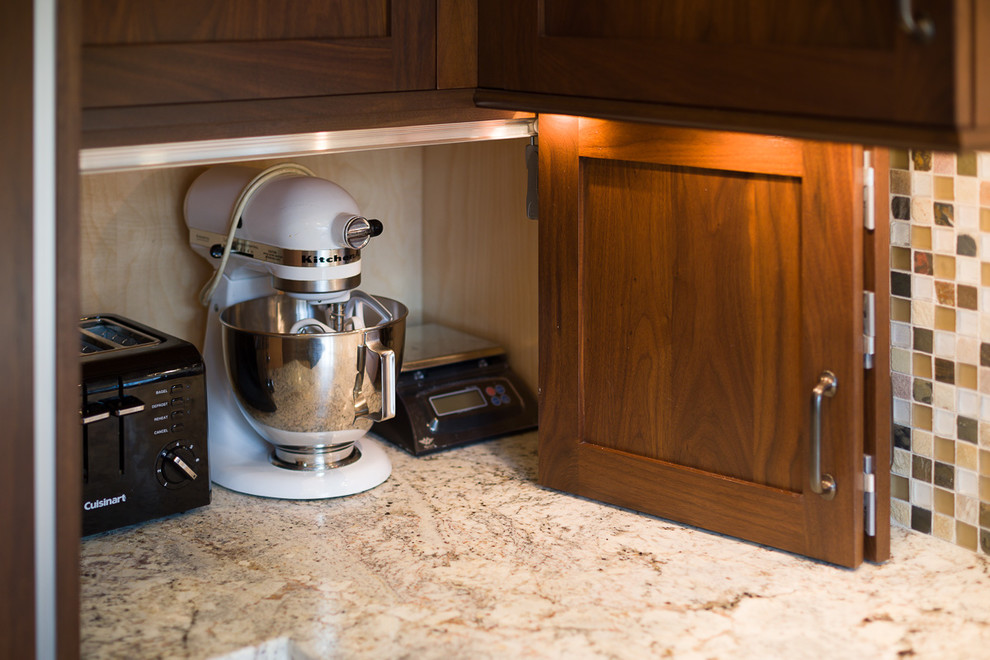 Amerikansk inredning av ett avskilt, litet u-kök, med en undermonterad diskho, skåp i shakerstil, bruna skåp, granitbänkskiva, flerfärgad stänkskydd, stänkskydd i mosaik, integrerade vitvaror, mellanmörkt trägolv och en halv köksö