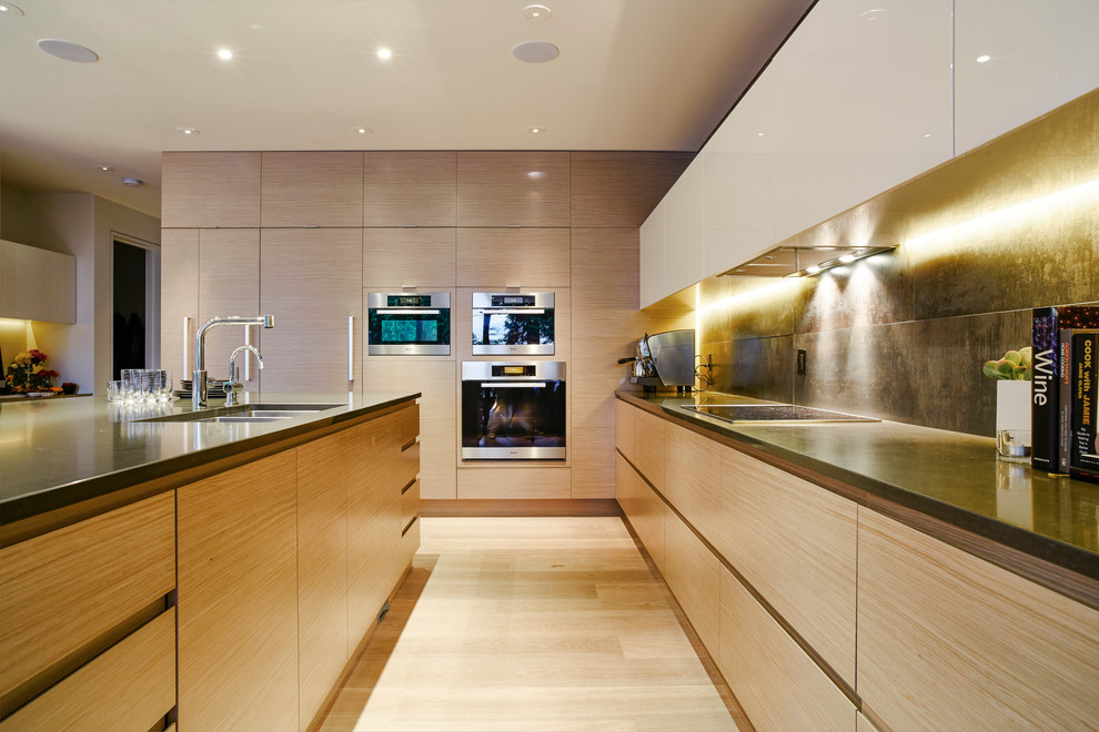 This is an example of a large contemporary l-shaped kitchen/diner in Vancouver with a submerged sink, flat-panel cabinets, light wood cabinets, engineered stone countertops, metallic splashback, porcelain splashback, stainless steel appliances, light hardwood flooring and an island.