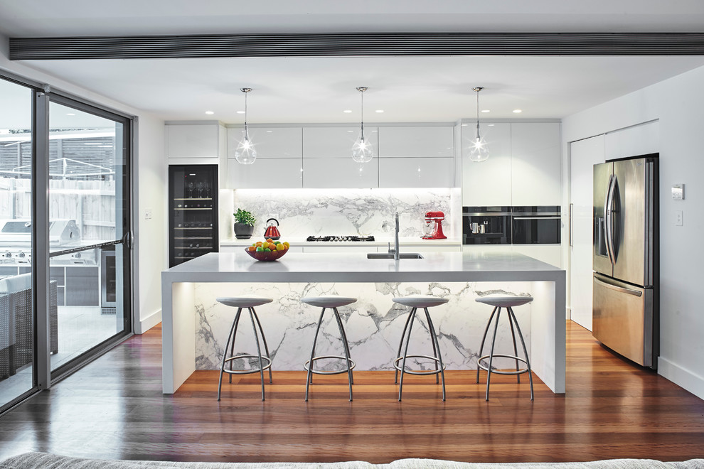 Offene Moderne Küche in L-Form mit Unterbauwaschbecken, flächenbündigen Schrankfronten, weißen Schränken, Küchengeräten aus Edelstahl, braunem Holzboden, Kücheninsel und Rückwand aus Marmor in Sydney